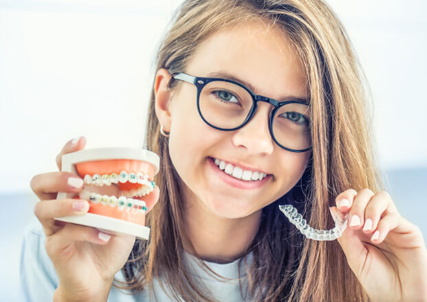 smiling girl holding up a model of teeth with braces and a clear aligner