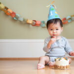 Asian baby wears a blue party hat and eats a smash cake at his first birthday