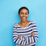 Black woman in a striped shirt smiles while standing against a sky blue wall