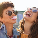 2 young women with sunglasses smile while one blows a gum bubble